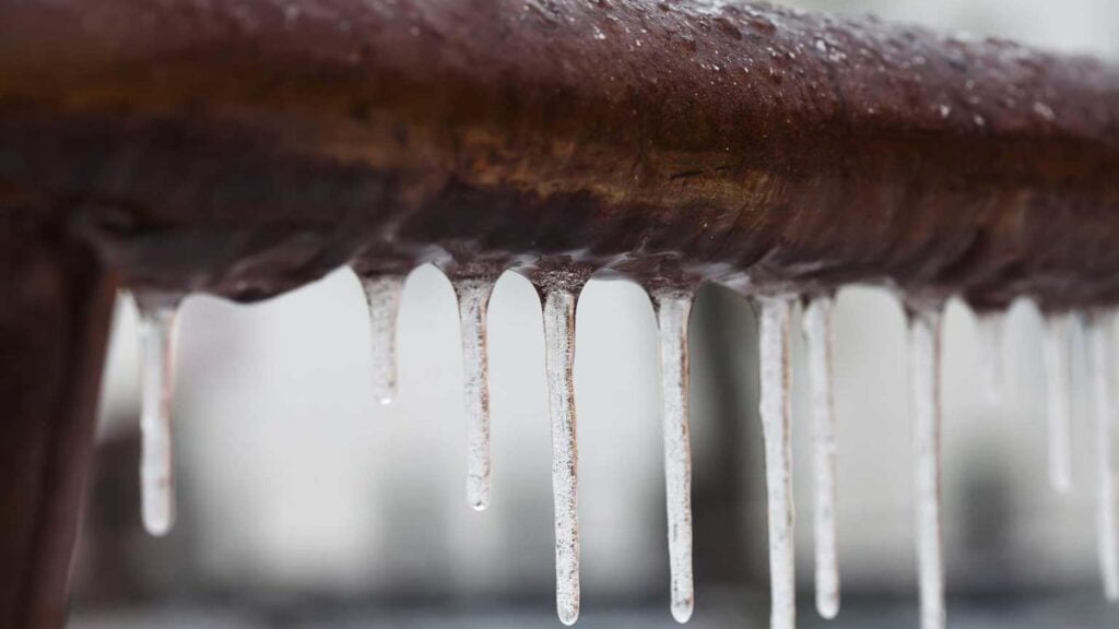 Icicles hanging from a frozen metal pipe covered in ice and frost during winter weather conditions.