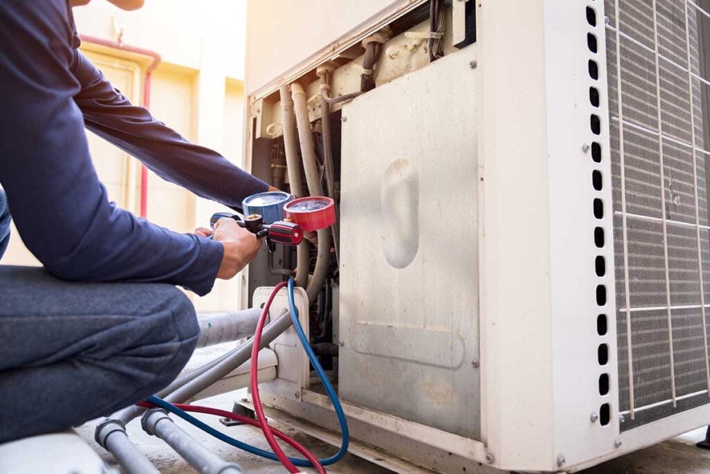 Technician holding pressure gauges with red and blue hoses connected to an air conditioning unit outdoor condenser.