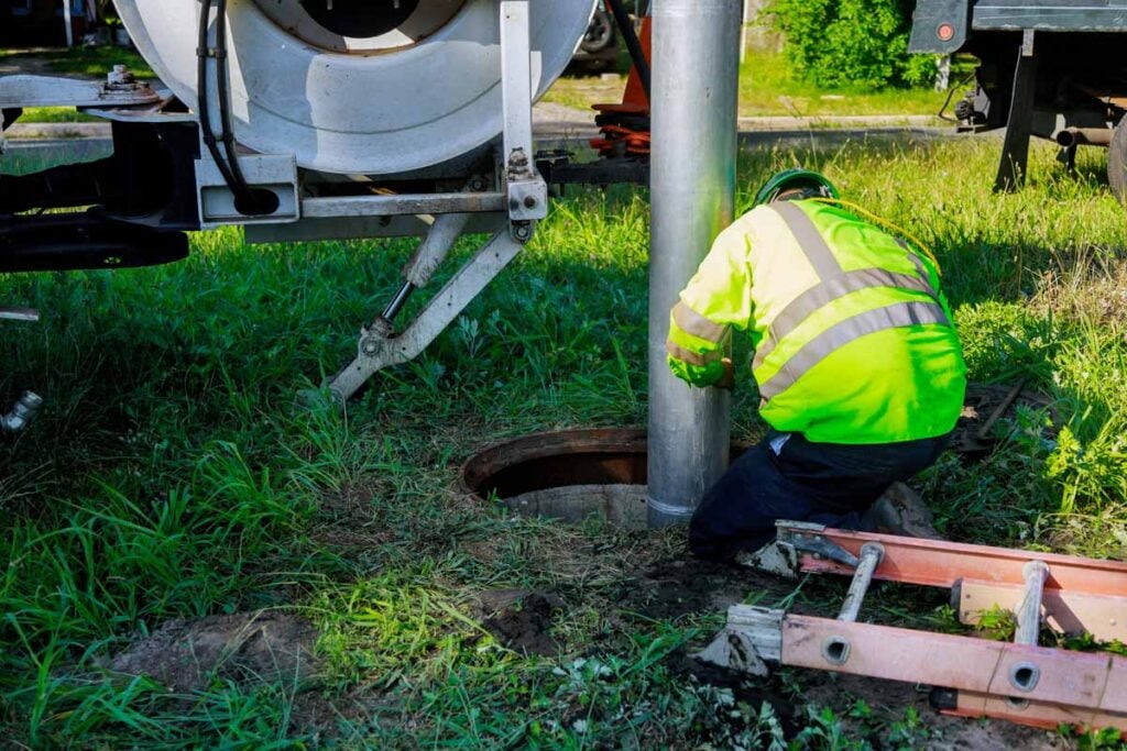 Utility worker in bright yellow safety vest inspecting an open sewer manhole with concrete pipe visible underground.