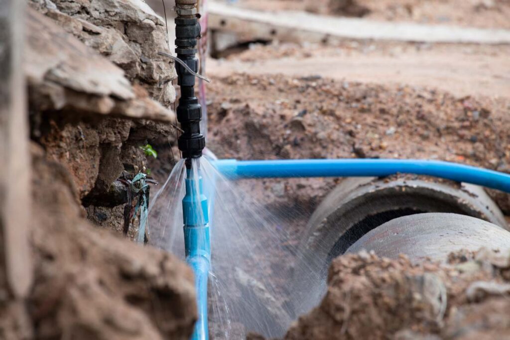 Blue drip irrigation hose spraying water from a black connector attached to a rough concrete or stone wall in an arid outdoor setting.