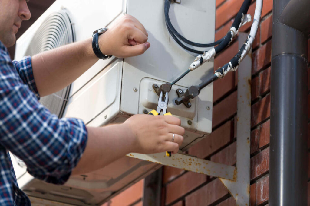 HVAC technician connecting refrigerant lines to an outdoor air conditioning unit mounted on a brick wall.