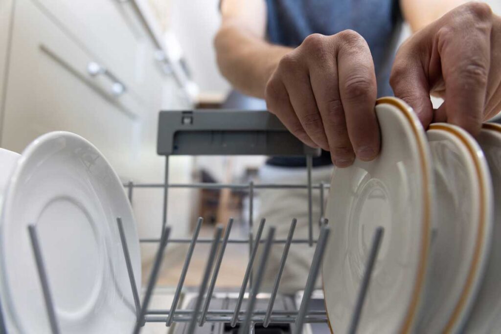 Person placing clean dishes with gold trim into the upper rack of an open dishwasher during loading.