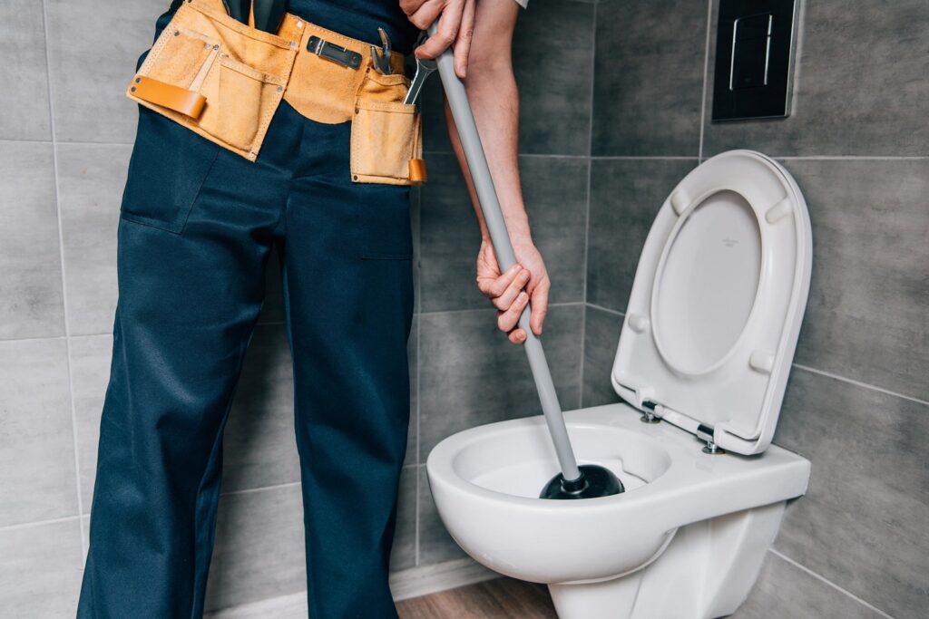 Professional plumber in work apron and dark pants uses a plunger to unclog a white toilet in a modern bathroom with gray tile walls.