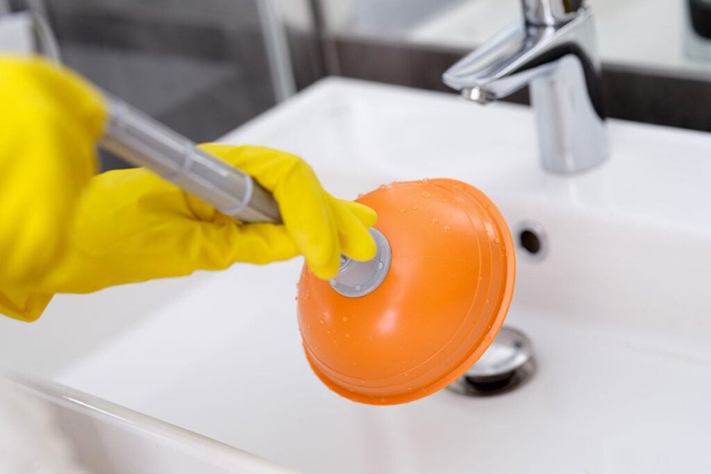 Person wearing yellow gloves uses a plunger to clear an orange suction cup from a white bathroom sink drain.