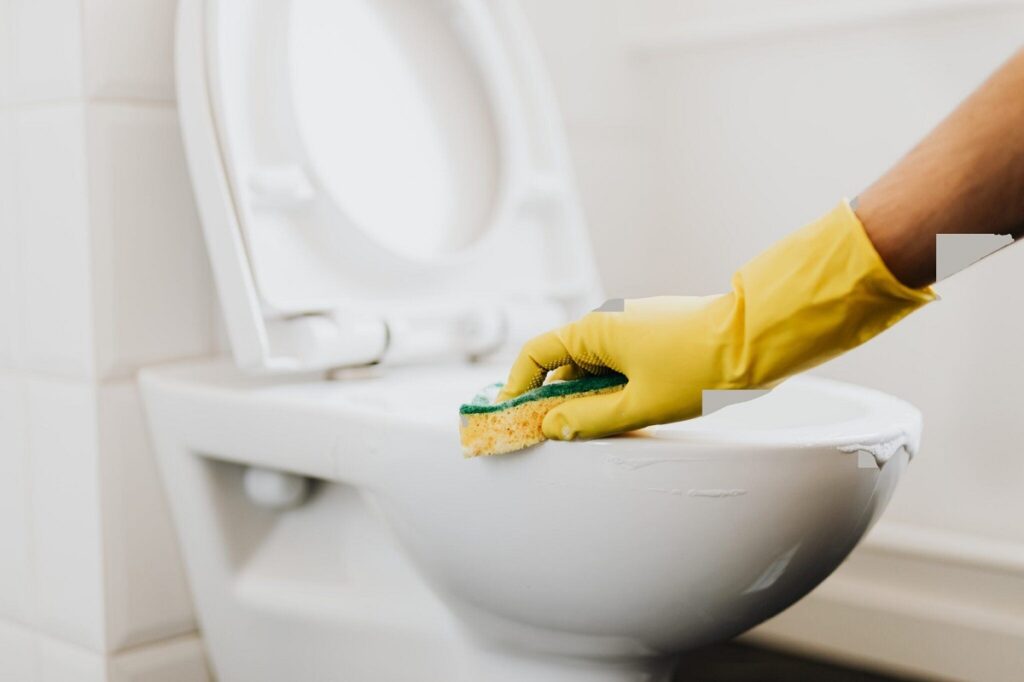 Person wearing yellow rubber gloves scrubbing inside a white toilet bowl with a yellow and green sponge.