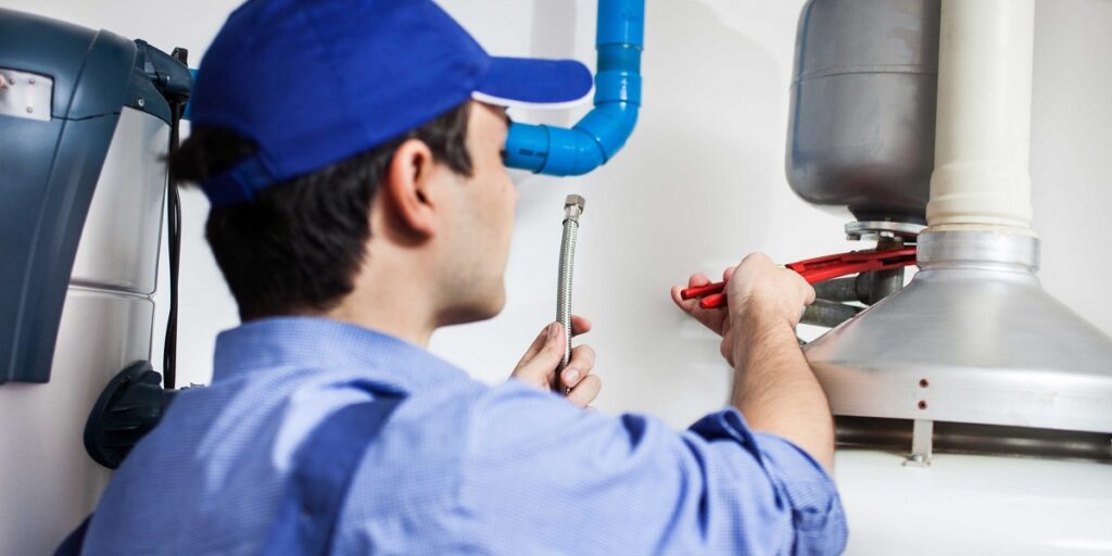 Professional plumber in blue uniform and cap working on hot water tank installation with wrench and flexible connector pipe.