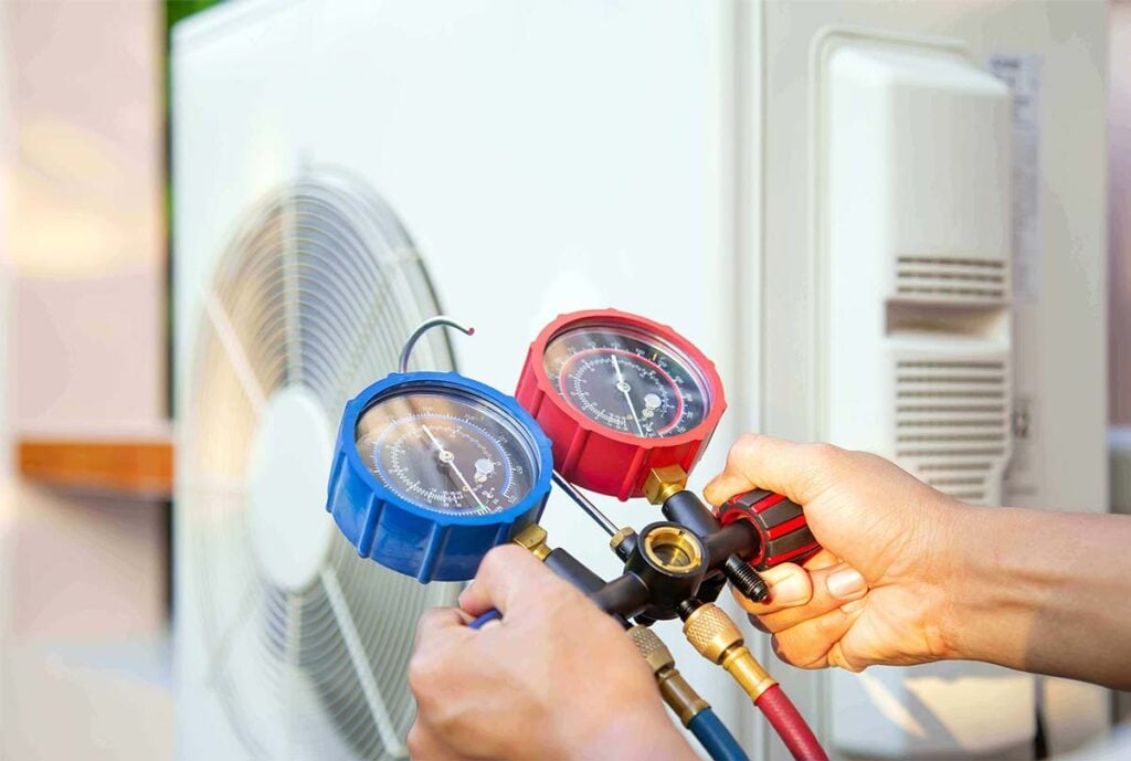 Technician holding blue and red pressure gauges connected to refrigerant lines while servicing an air conditioning unit with visible frost buildup.