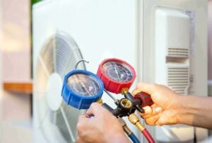 Technician holding blue and red pressure gauges connected to refrigerant lines while servicing an air conditioning unit with visible frost buildup.