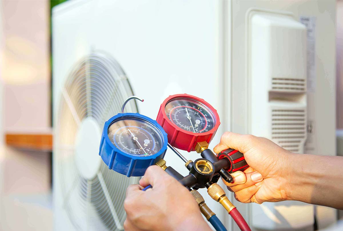 Technician holding blue and red pressure gauges connected to refrigerant lines while servicing an air conditioning unit with visible frost buildup.