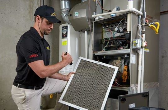 Technician in black shirt holding pleated air filter while servicing residential furnace unit mounted on wall.