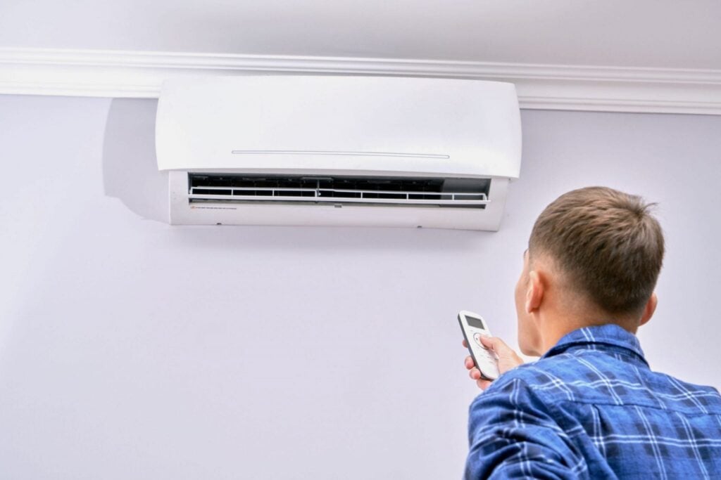 Man in blue plaid shirt holding remote control while standing below a white wall-mounted ductless air conditioning unit.