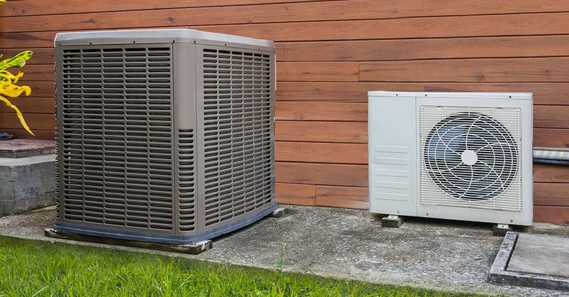 Two outdoor HVAC units mounted against a wooden wall next to grass, showing a larger gray condenser unit and smaller white heat pump unit.