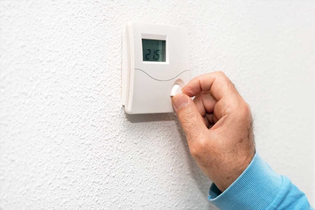A person's hand adjusting a white digital thermostat mounted on a textured wall, showing a temperature reading of 27.5 degrees.