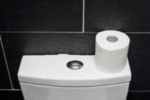 White toilet tank with flush button and roll of toilet paper on top against black tiled bathroom wall.