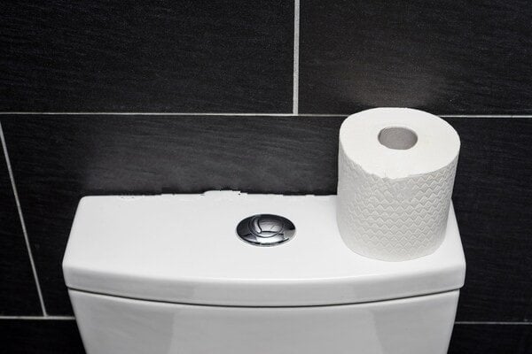 White toilet tank with flush button and roll of toilet paper on top against black tiled bathroom wall.