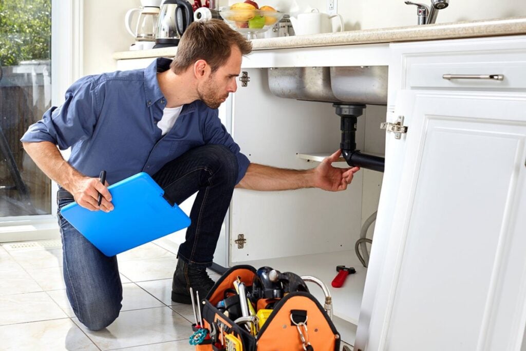 Professional plumber in blue shirt inspecting under-sink plumbing with clipboard and toolbox nearby.