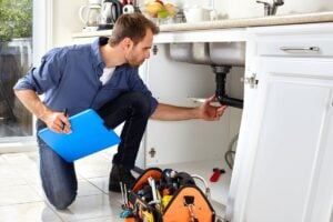 Professional plumber in blue shirt inspecting under-sink plumbing with clipboard and toolbox nearby.