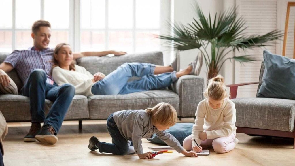 Parents sitting on gray couch watching children play with toys and coloring on wooden floor in bright living room.