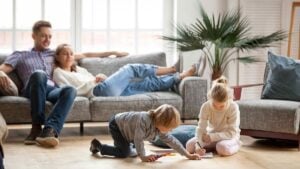 Parents sitting on gray couch watching children play with toys and coloring on wooden floor in bright living room.