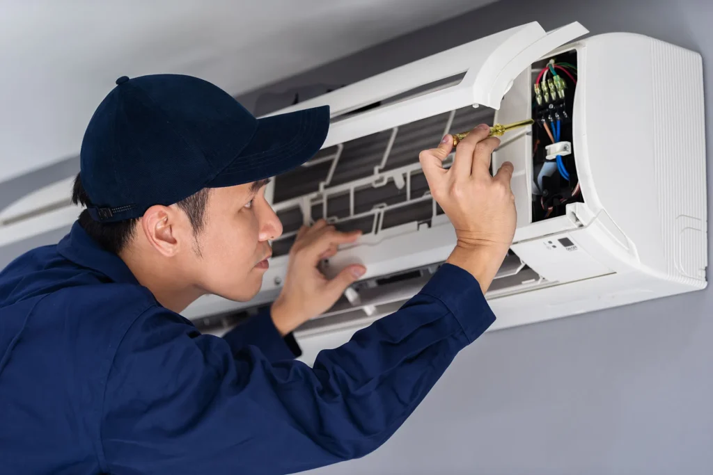 Technician in dark blue uniform and cap installing or servicing an indoor air conditioning unit, working with wiring and components.