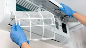 Person wearing blue gloves holding a white mesh air filter in front of an air conditioning unit.