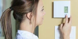 Woman in white shirt adjusting a digital wall thermostat displaying temperature on a yellow wall.