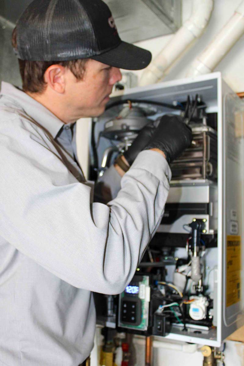 Technician in black cap and work gloves inspecting internal components of residential HVAC furnace unit during maintenance service.
