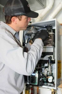 HVAC technician wearing black cap and gloves performing maintenance on an open furnace unit.