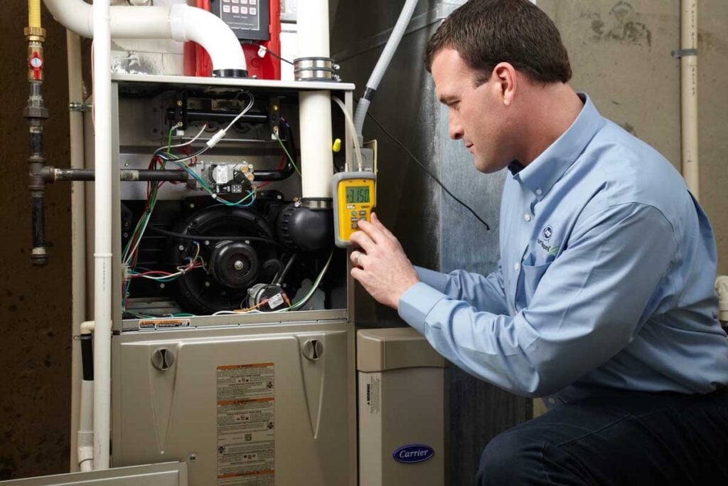 Professional technician in blue shirt examines interior components of Carrier furnace using yellow diagnostic multimeter tool.