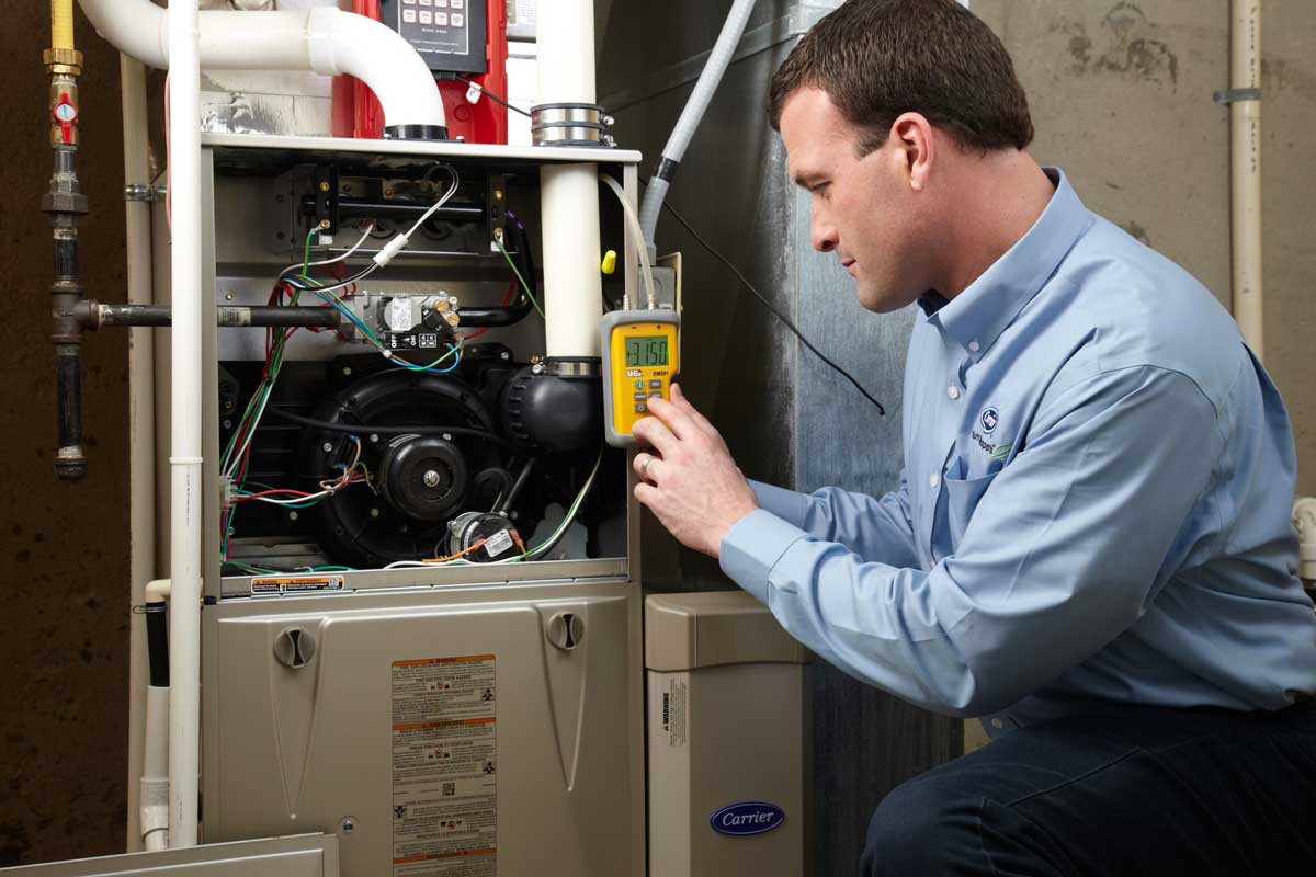 Professional technician in blue shirt examines interior components of Carrier furnace using yellow diagnostic multimeter tool.