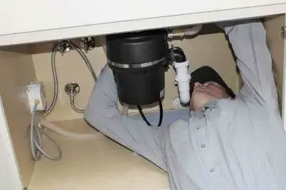 Plumber in white shirt working under kitchen sink to install or repair black drain pipe and plumbing connections.