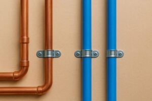 Copper and blue PEX pipes mounted on a beige wall with metal U-bracket clamps securing them in place.
