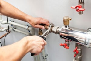 Plumber's hands tightening metal pipe fittings with a wrench, with red valve handles and white plastic pipes visible in background.