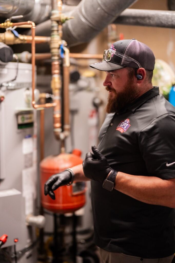 Bearded technician wearing black gloves and cap inspects HVAC equipment and copper piping in mechanical room.