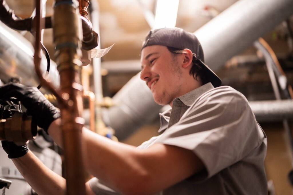 Male plumber wearing a cap and gray shirt working on copper pipes and fittings with tools in an industrial setting.