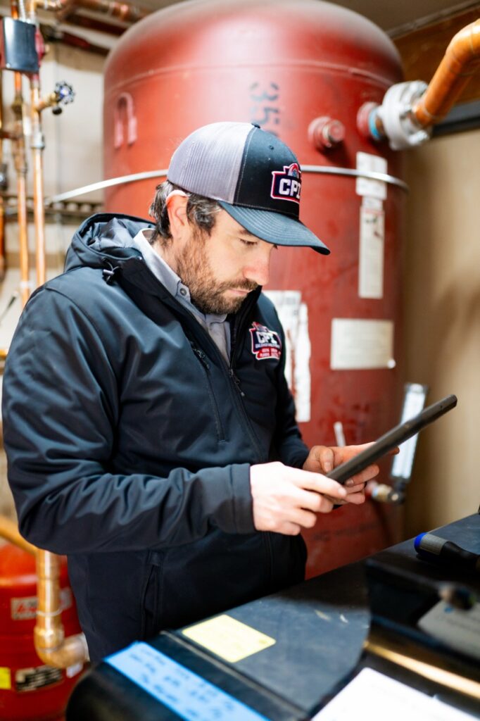 Technician wearing a cap and dark jacket examines a tablet near red industrial HVAC equipment in a mechanical room.