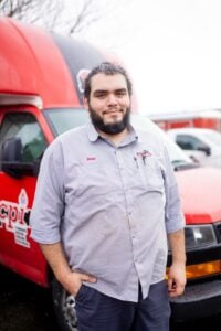 Male worker wearing light blue button-up shirt with name tag standing in front of red service vehicles.