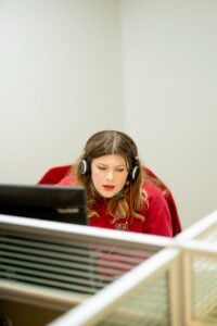 Woman wearing red shirt and headphones with microphone sits at desk facing monitor while working.