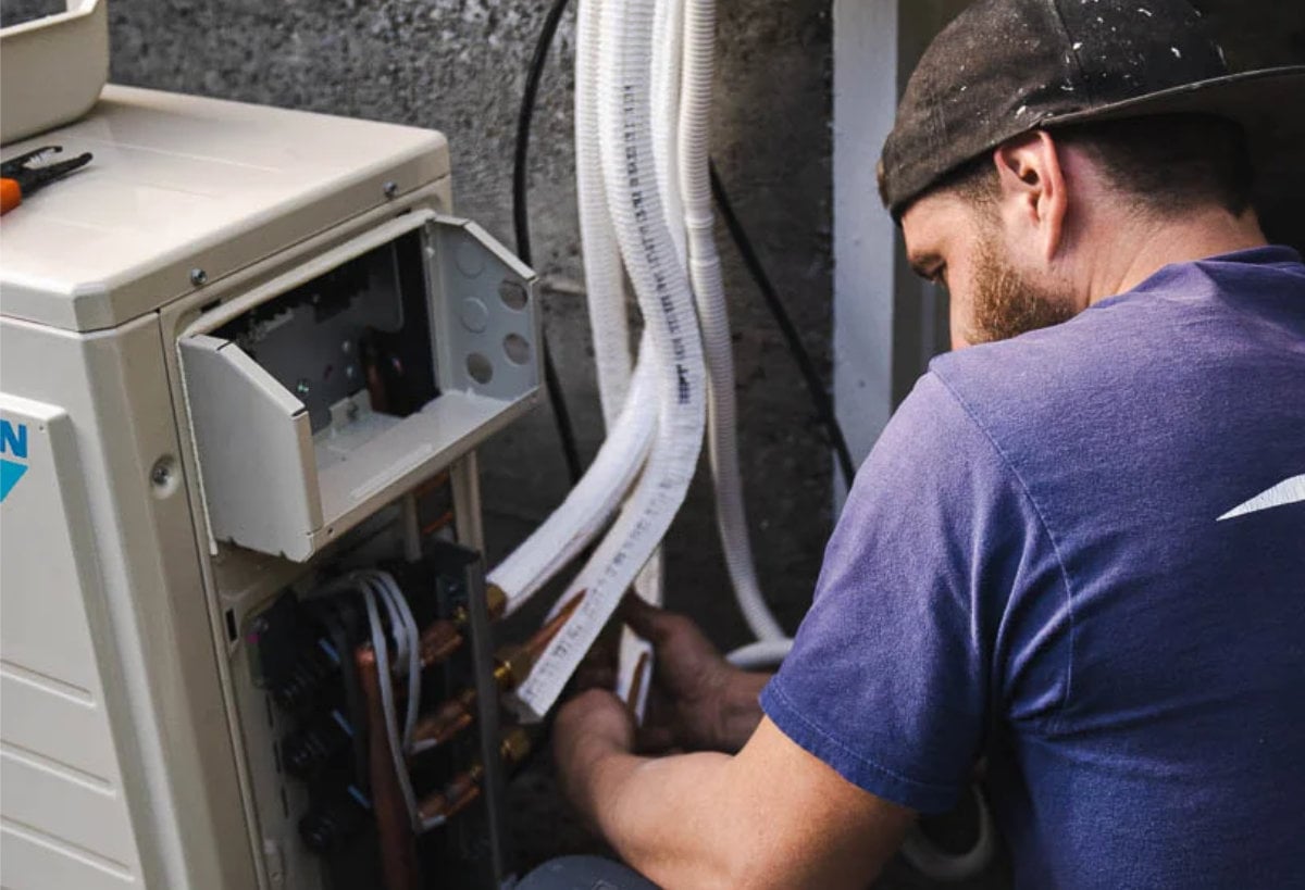 Technician in blue shirt and cap performing maintenance on an open heat pump unit with visible wiring and components.