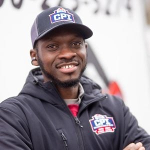 Smiling man wearing a dark CPI baseball cap and matching dark jacket with CPI logo over a red shirt.
