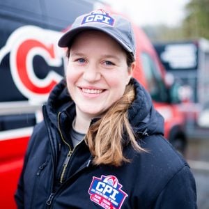 Smiling young woman wearing a navy CPT baseball cap and matching branded jacket standing in front of red vehicles.